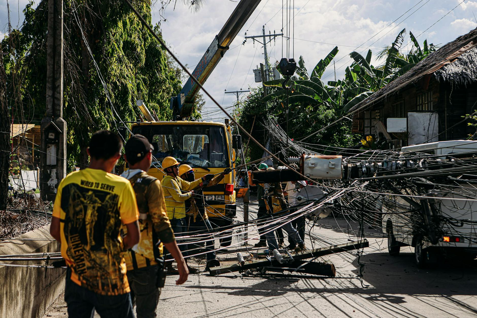Workers fixing damaged electrical lines on a street post-storm, surrounded by fallen cables and equipment.