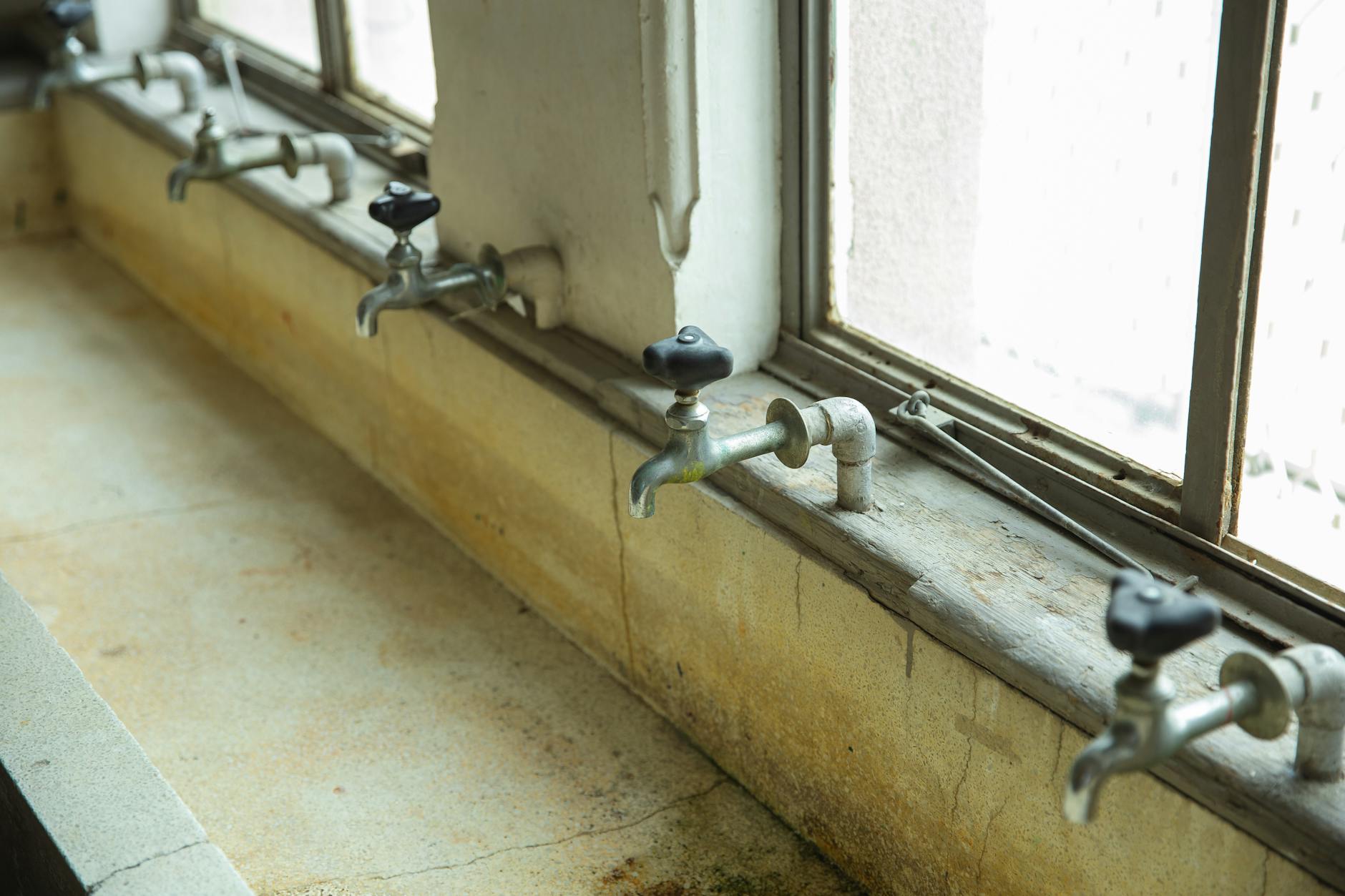From above of row of aged metal taps over dirty sink near shiny windows in sunlight