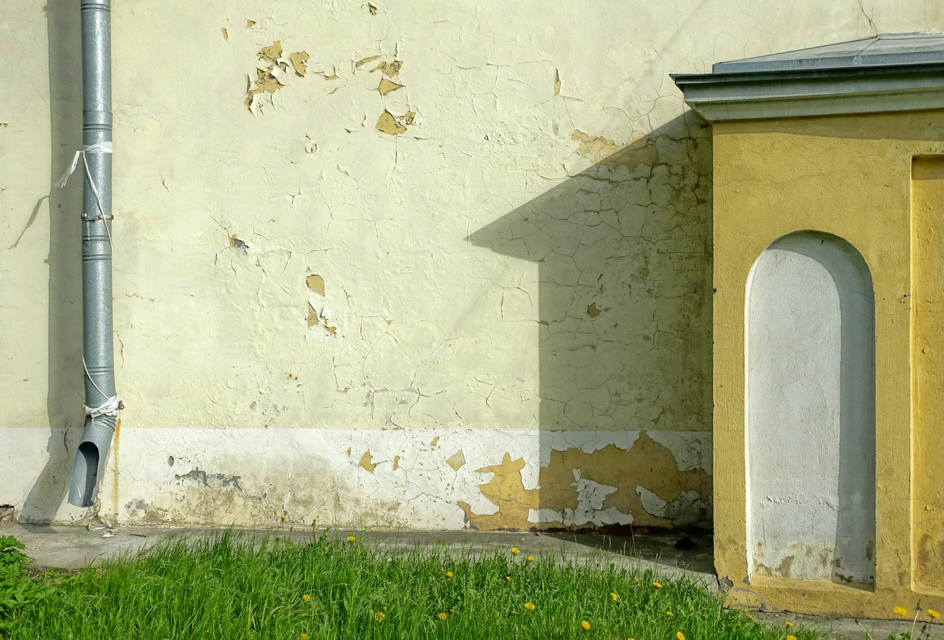 Detailed view of an old wall with cracked paint, downpipe, and grass at the base.