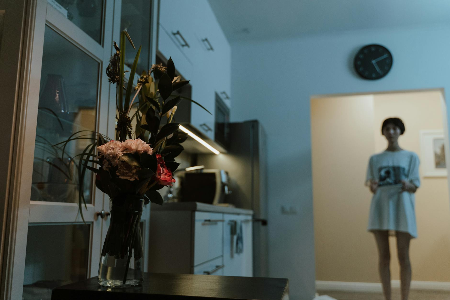 A modern kitchen interior featuring a glass vase of flowers with subtle lighting and a blurred figure.