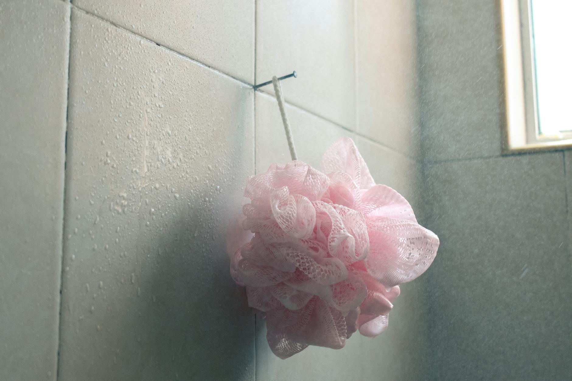 A pink loofah hangs in a tiled bathroom surrounded by water droplets.