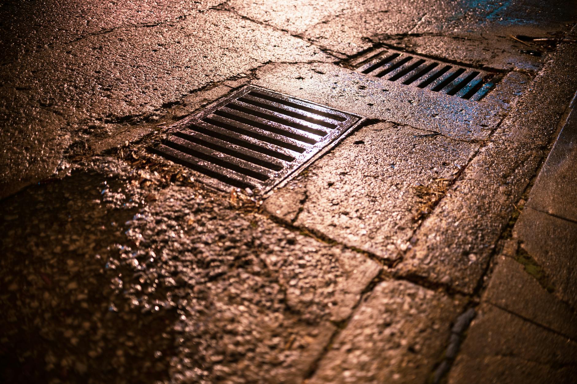 Nighttime shot of wet asphalt road with drainage grates reflecting light.