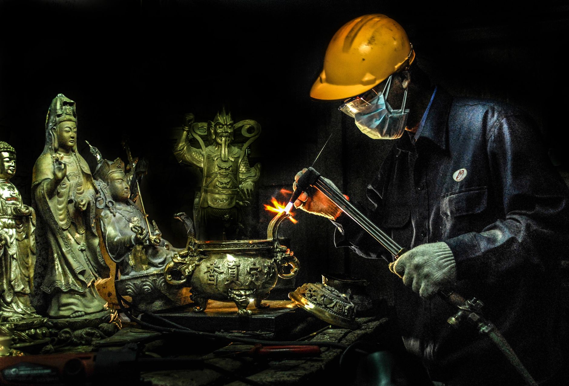 A craftsman wearing a helmet and mask welds religious statues in a dimly lit workshop.