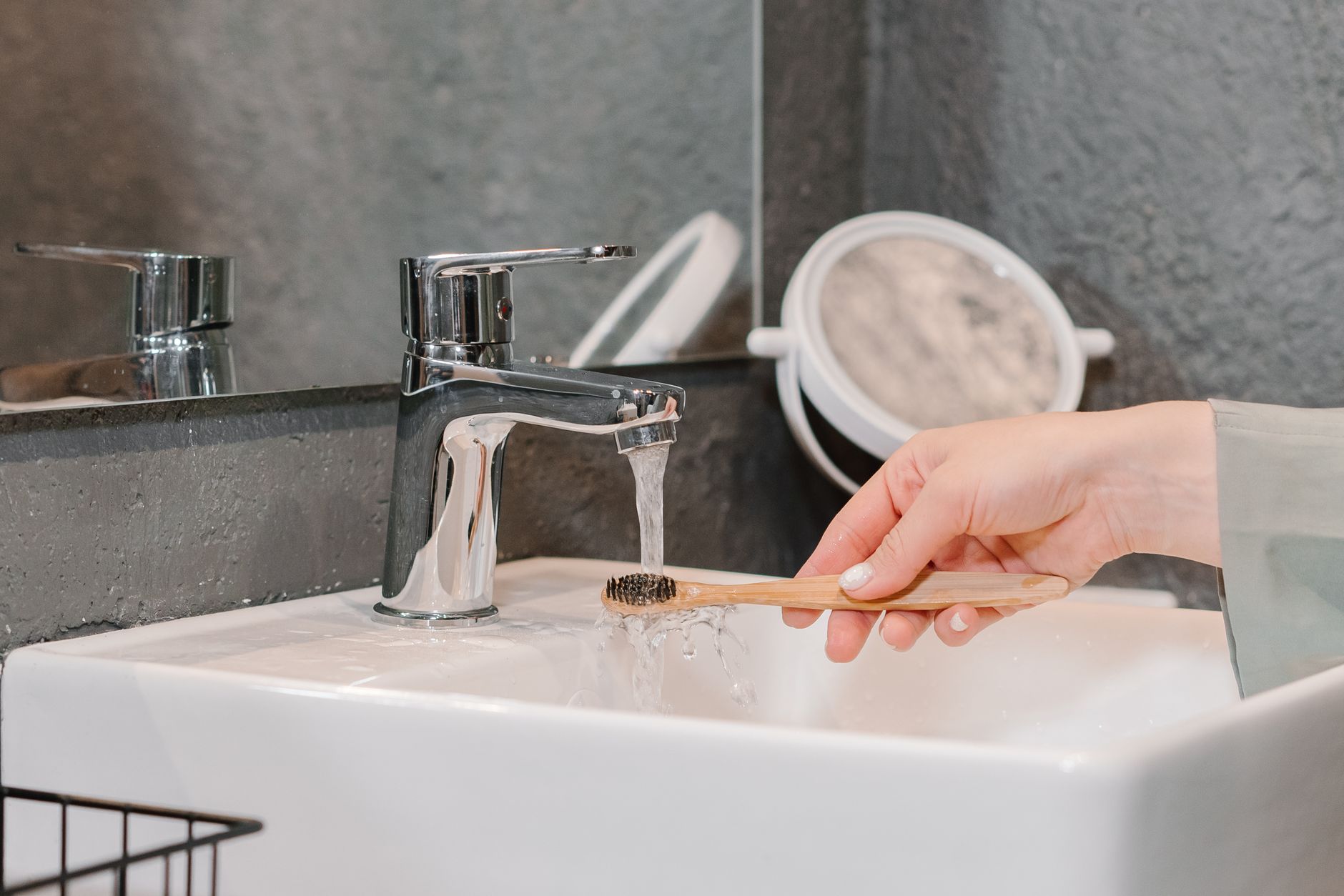 A hand rinsing a bamboo toothbrush under a tap in a modern bathroom sink.