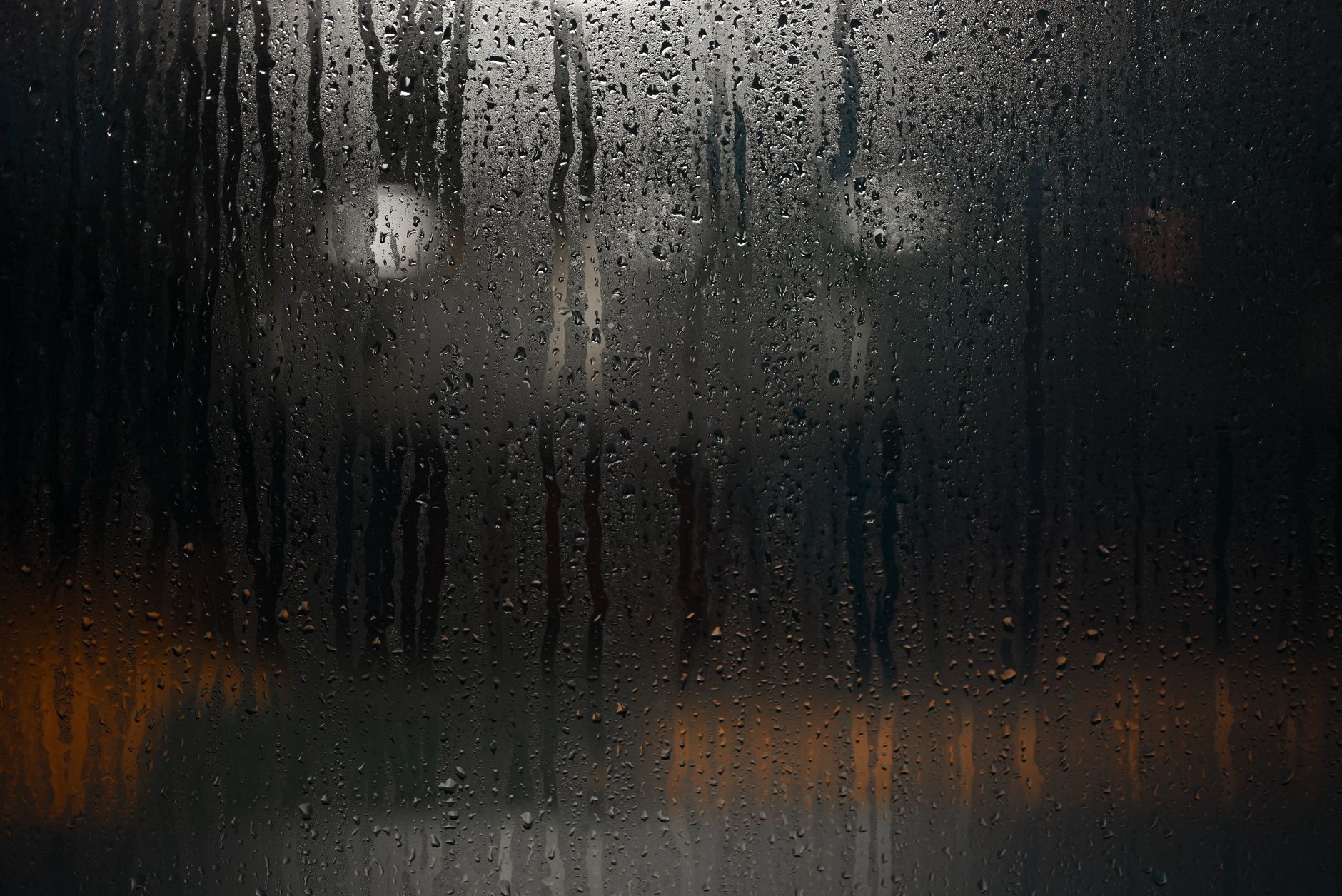 Rain-soaked window with droplets and reflections at night, creating a moody urban ambiance.
