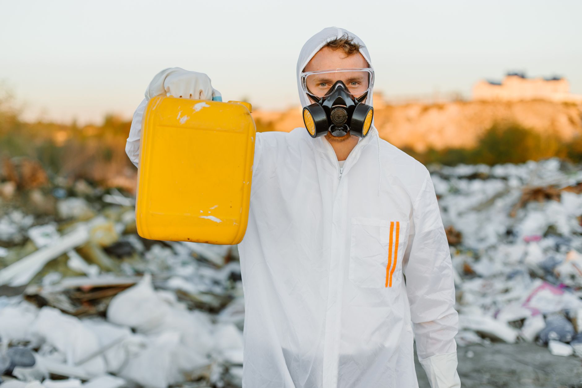 Worker in a protective suit holds a yellow container, monitoring pollution at a waste site.