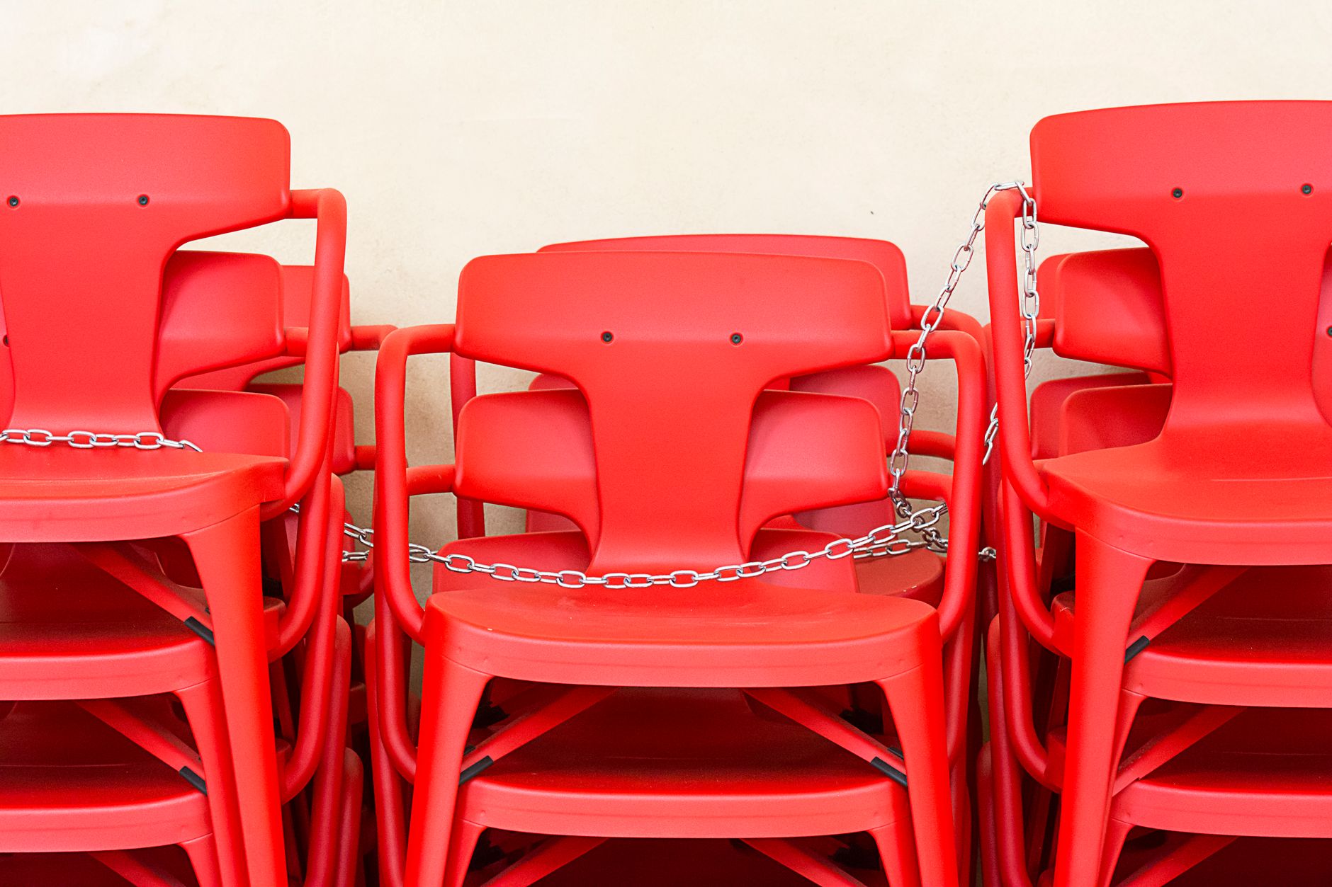 A stack of red plastic chairs secured with chains against a neutral background.