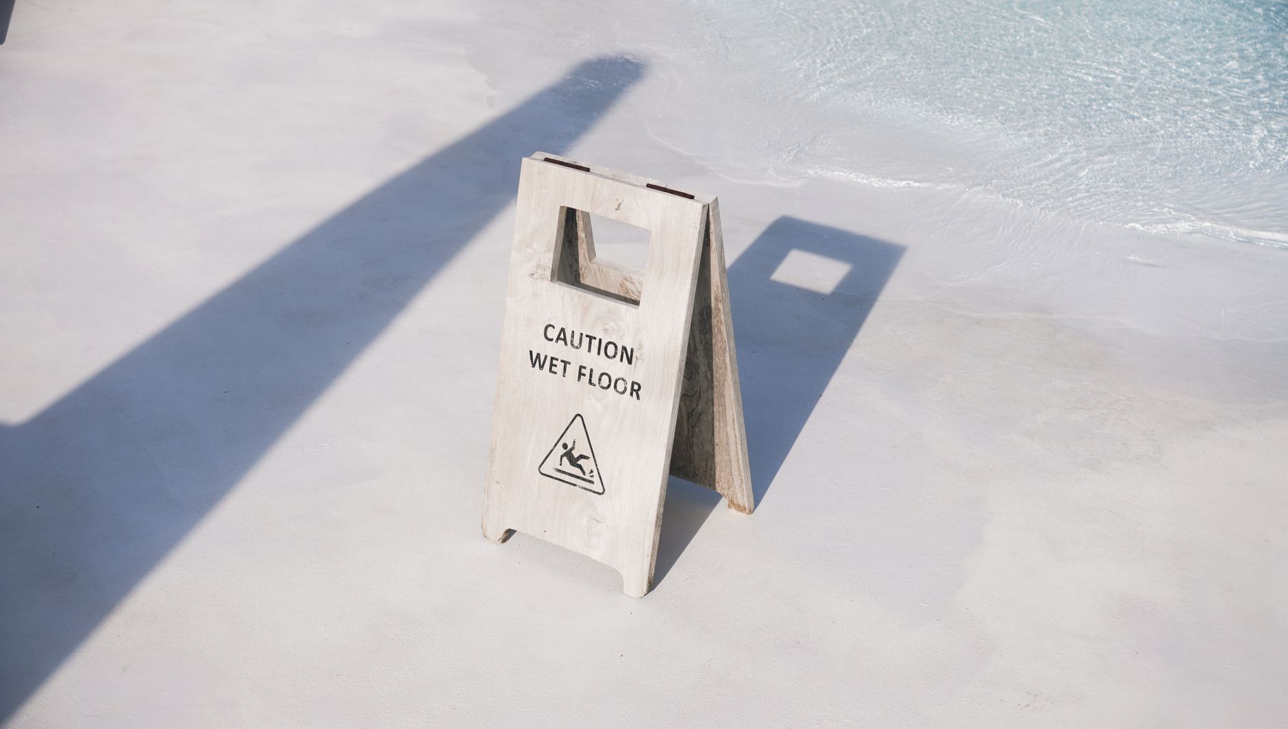 A caution wet floor sign placed near a poolside in bright daylight, casting shadows on the ground.