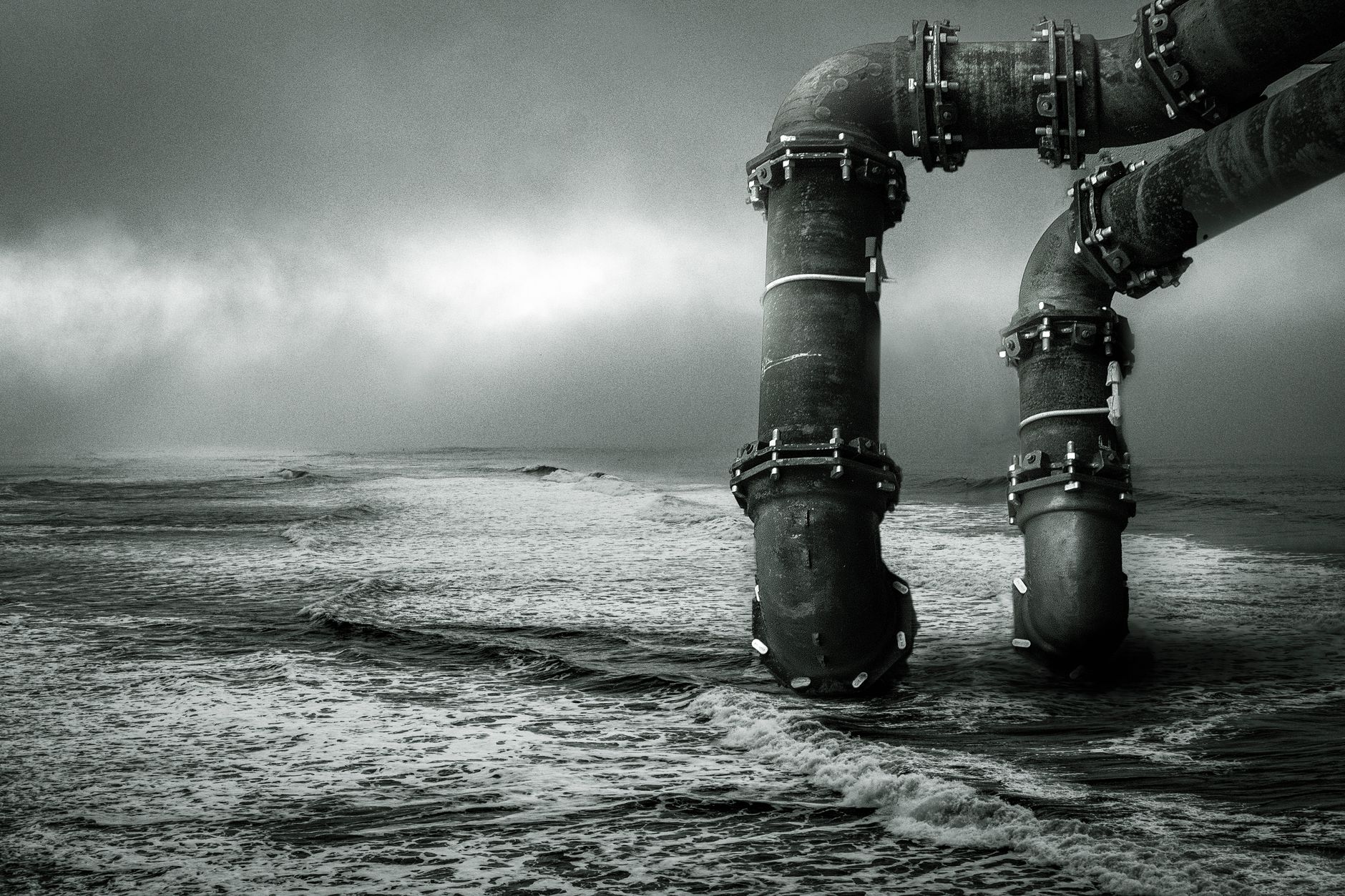 Monochrome image of large industrial pipes extending into the ocean under a dramatic sky.