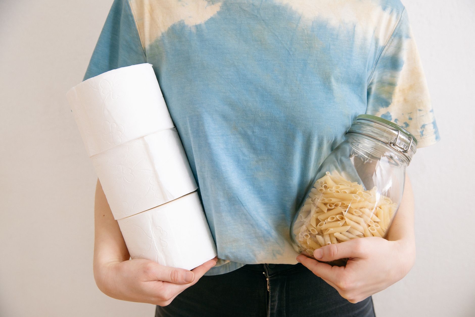 Woman in tie-dye shirt holding toilet paper rolls and pasta jar indoors.