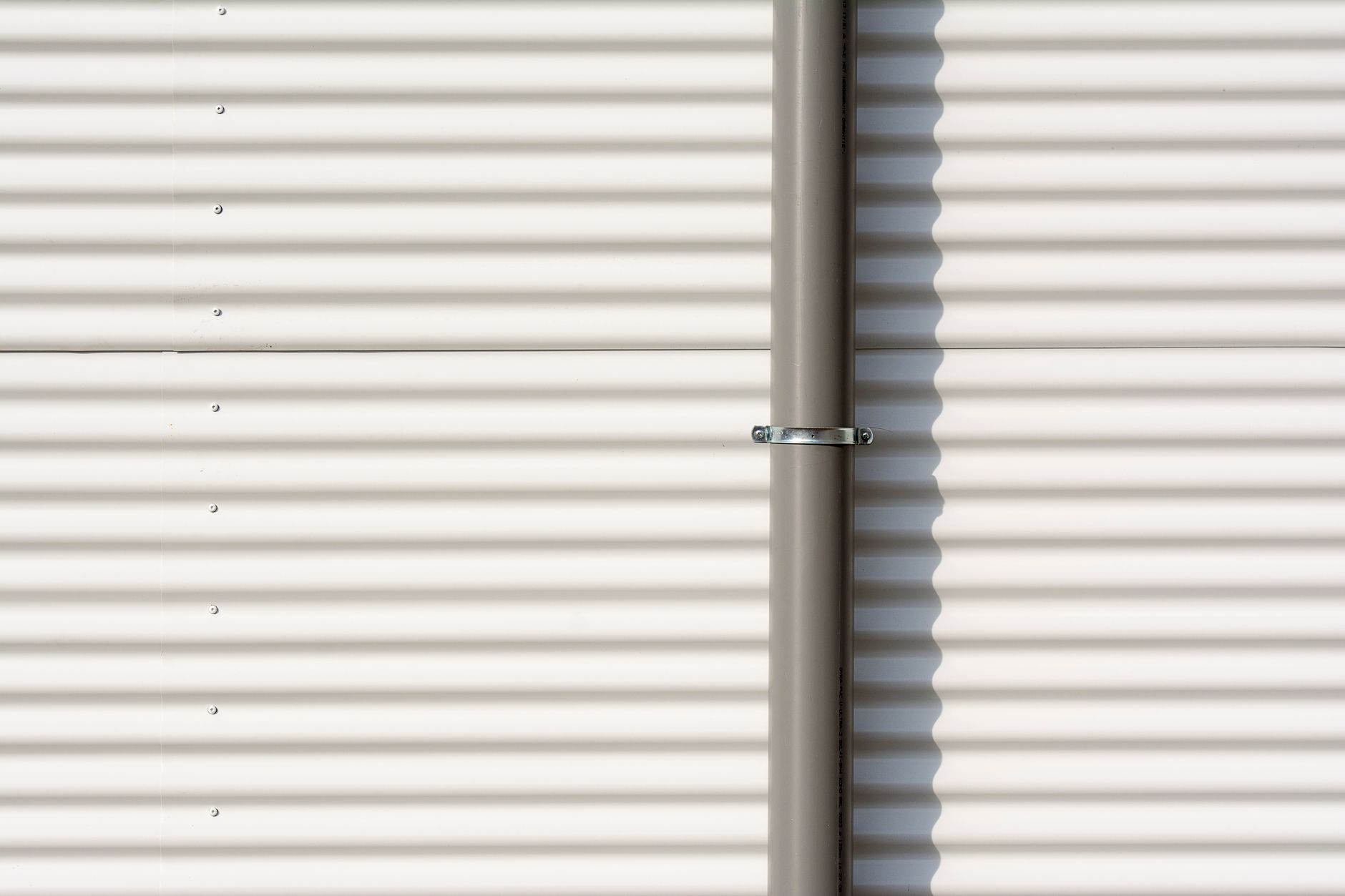 Close-up of a corrugated aluminum wall with a vertical drain pipe casting shadows.