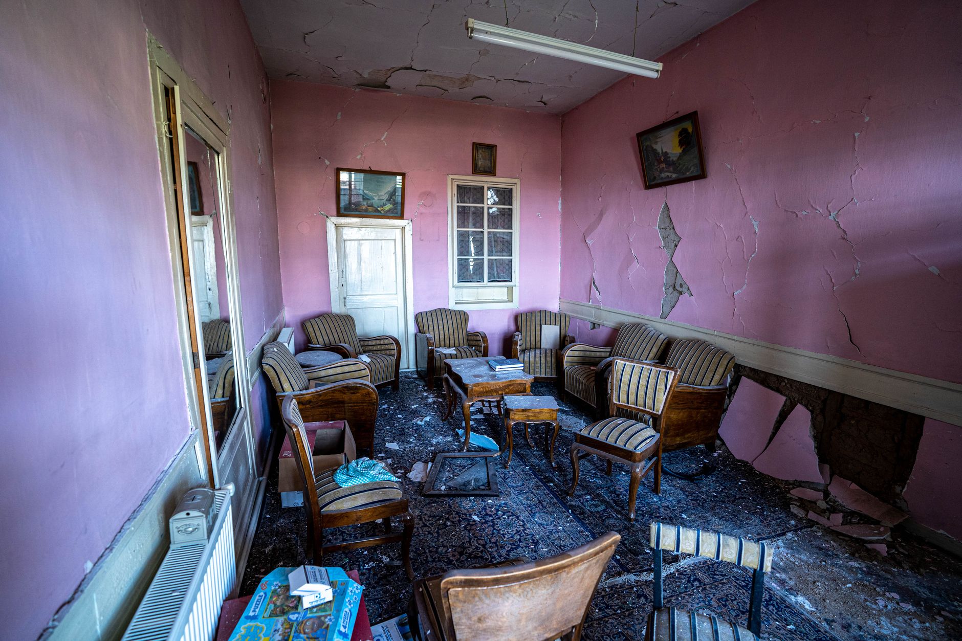 An abandoned living room with vintage furniture, peeling pink walls, and debris on the floor.