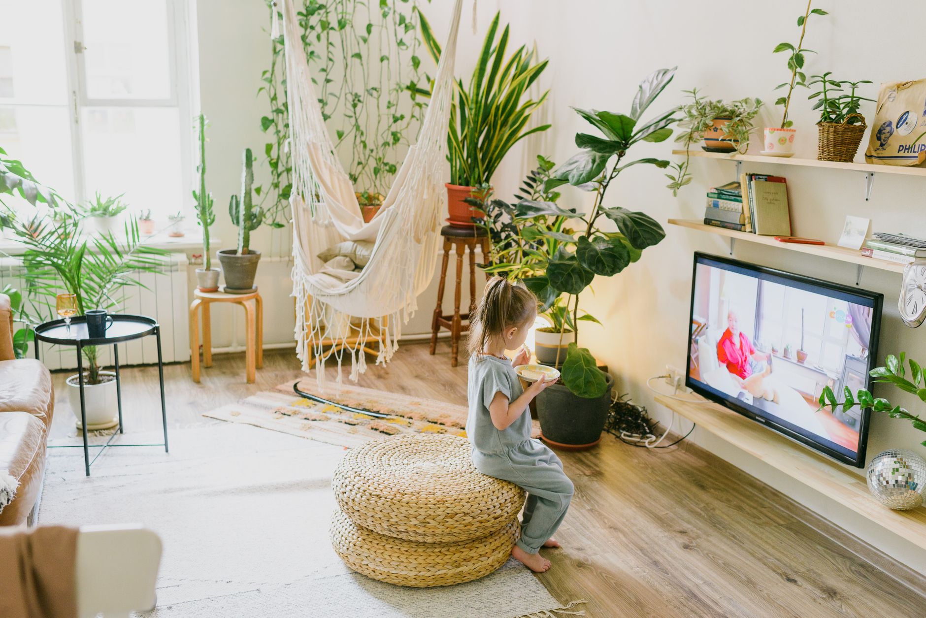 Child enjoying a snack in a cozy living room with lush plants and a hammock chair.