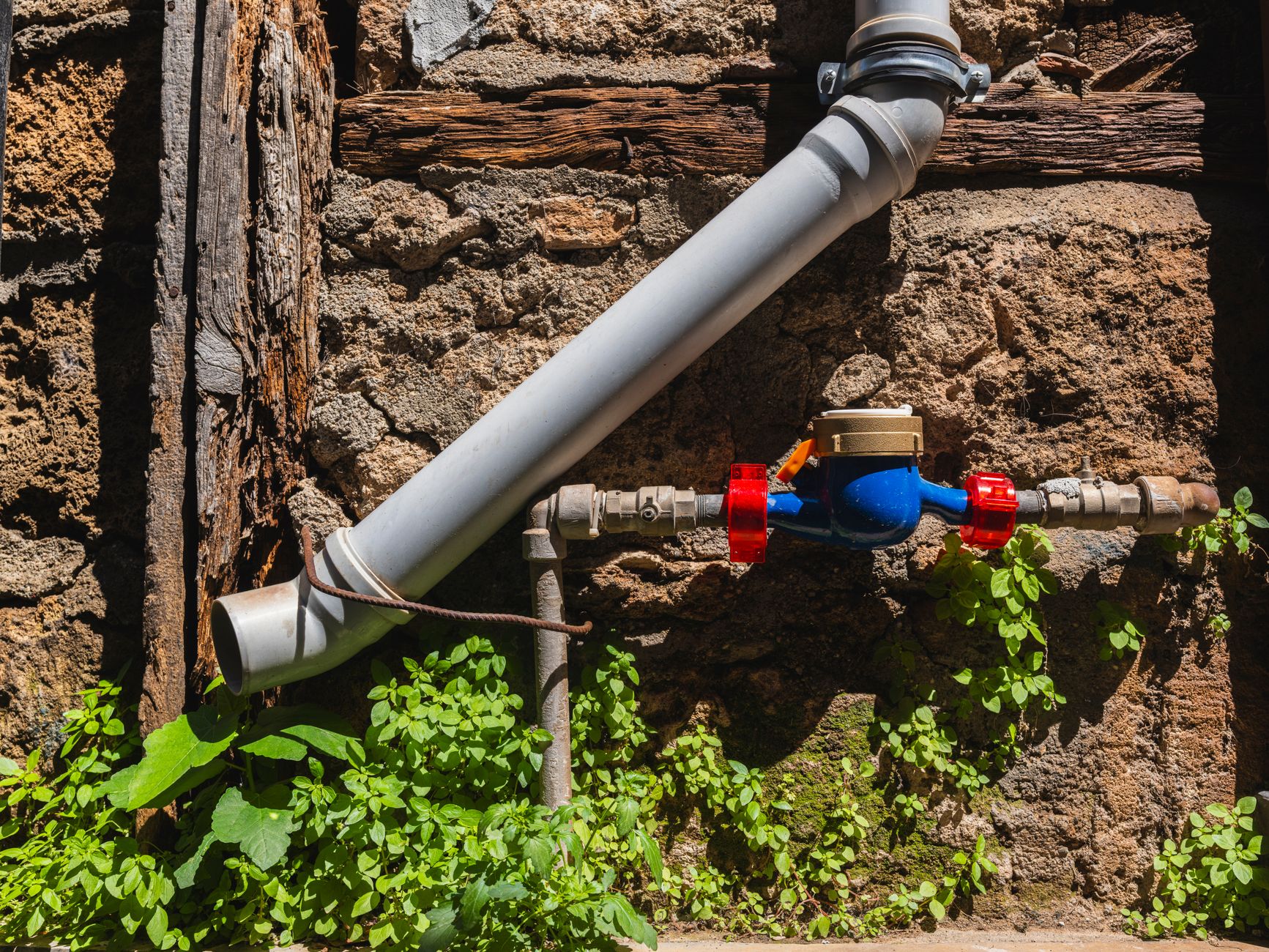 A weathered stone wall with a water pipe, meter, and greenery, depicting rustic charm.