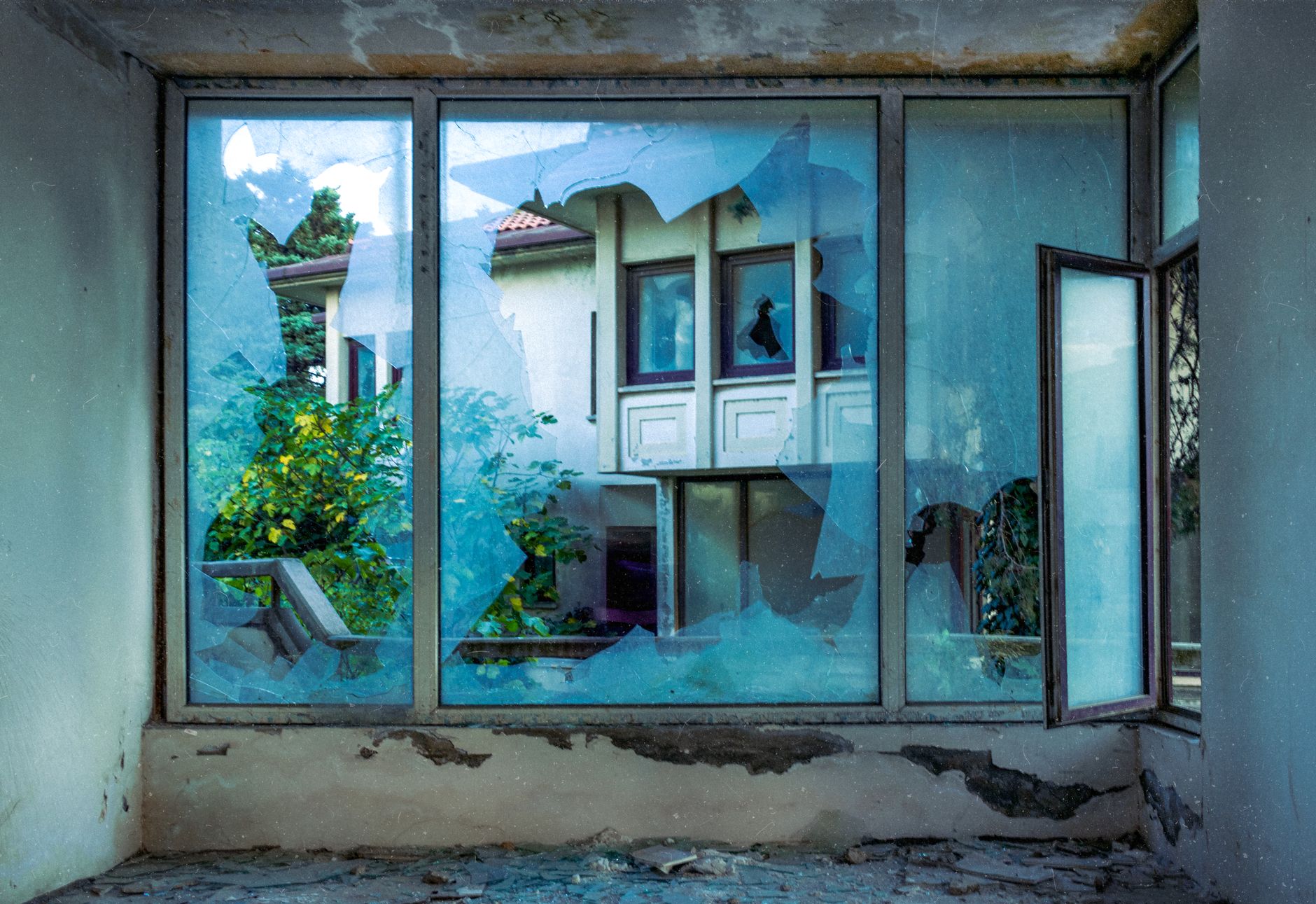 View through broken window of an empty house, showcasing decay and nature.