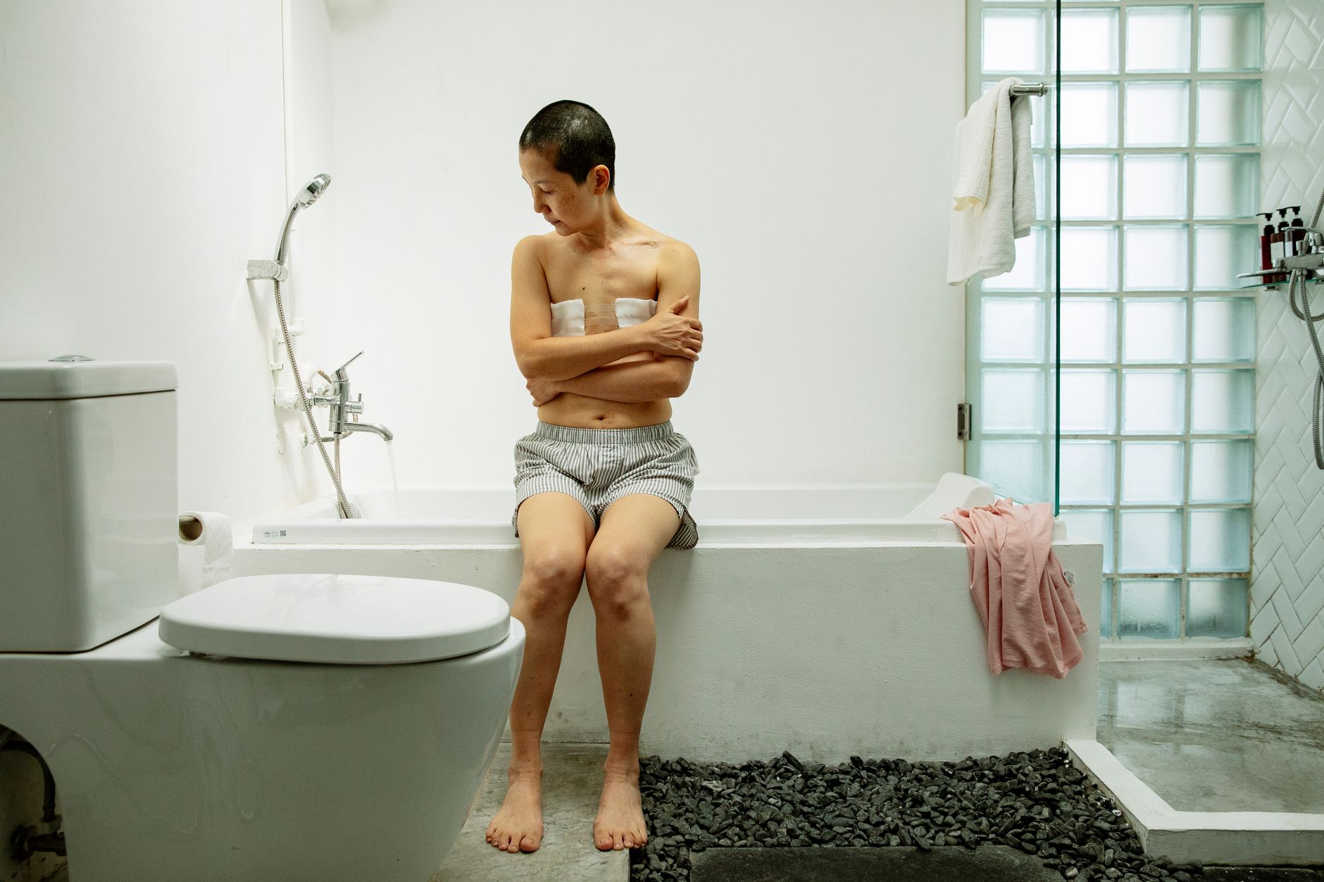 Full body of shirtless woman with short dark hair and sterile bandages after operation on breast sitting with crossed arms on bath in light bathroom