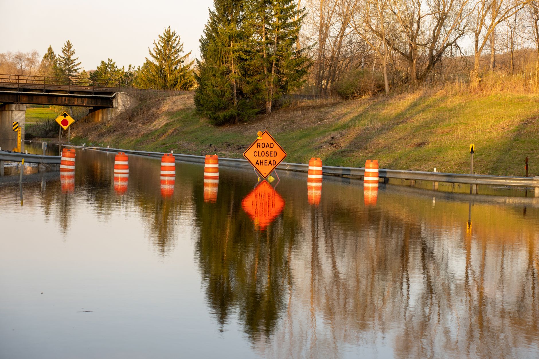 A flooded road with warning signs indicating 'Road Closed Ahead' due to water overflow.