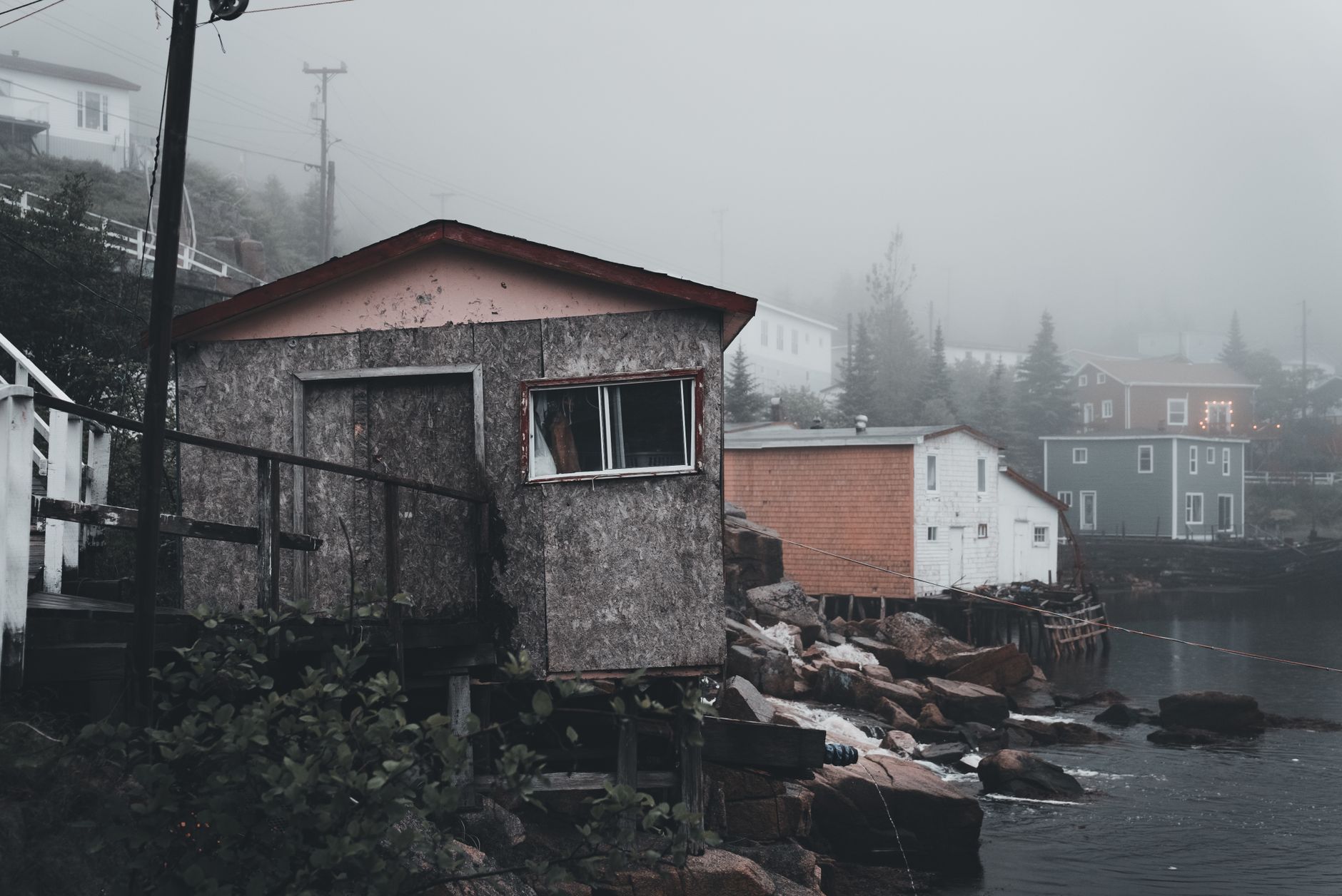 A foggy landscape of an abandoned coastal town featuring rustic houses by the water's edge.