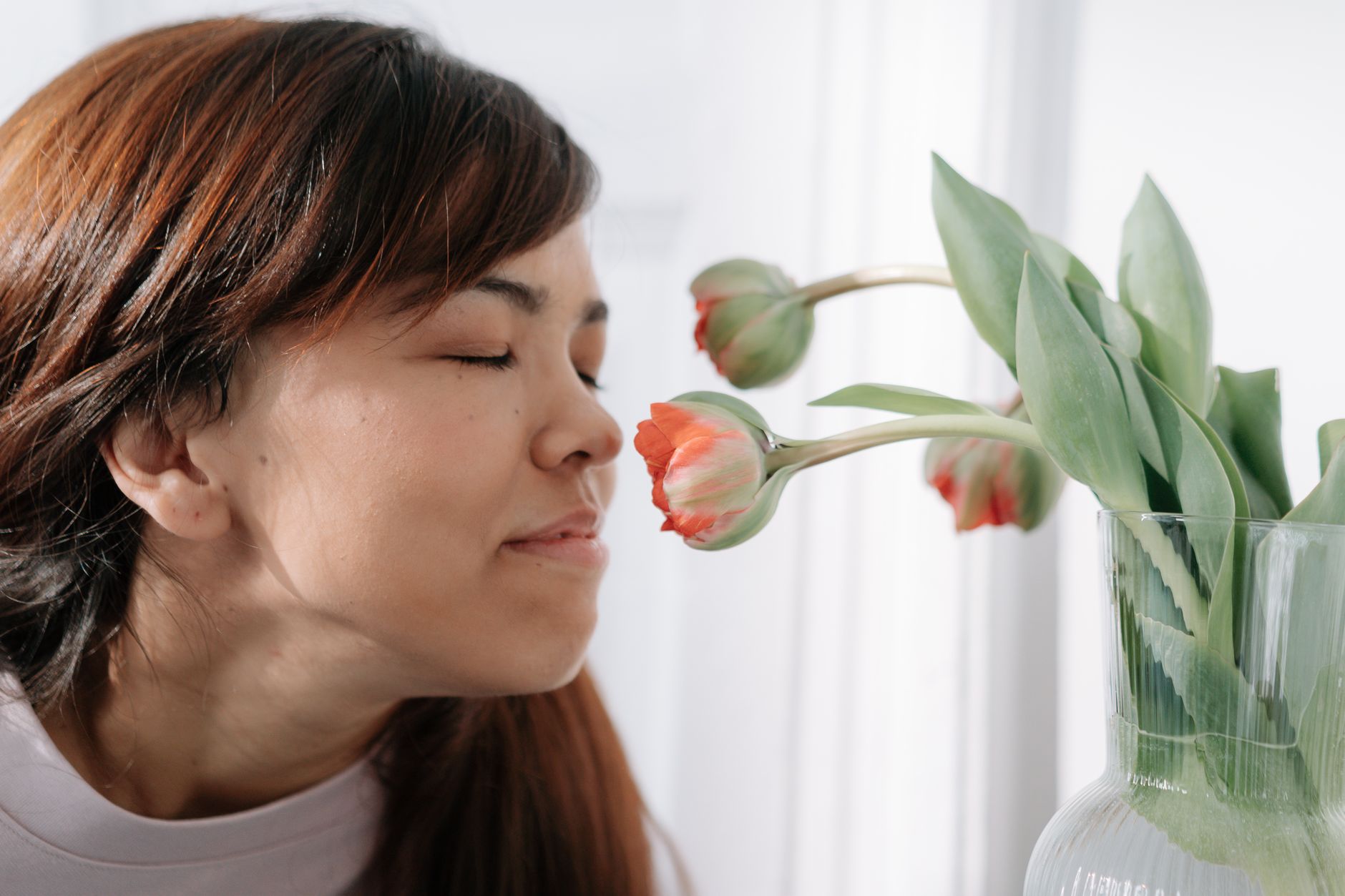 A woman with brown hair enjoys smelling tulips indoors, eyes closed in contentment.