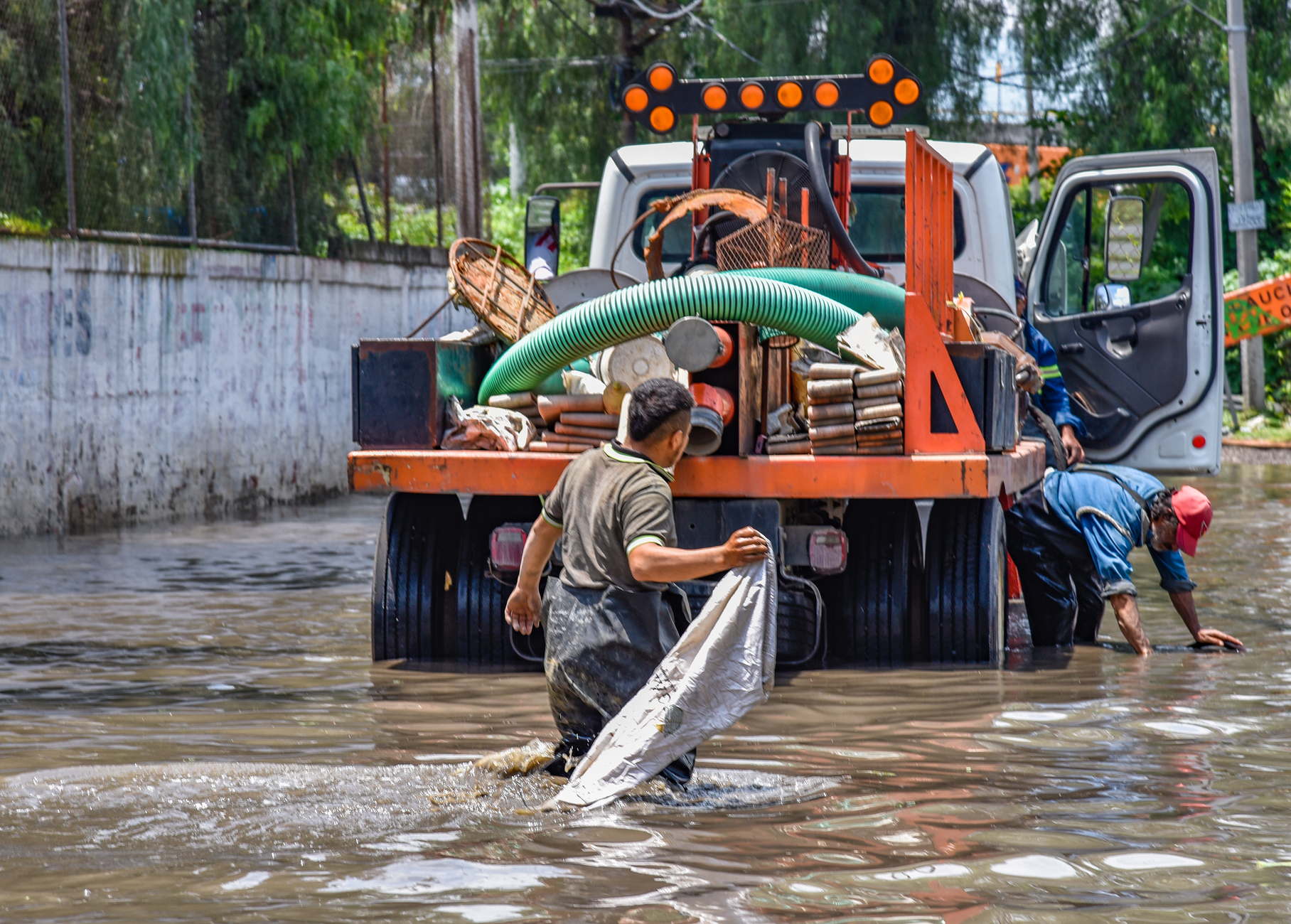 Workers wading through floodwaters near a truck, managing a city crisis after heavy rain.