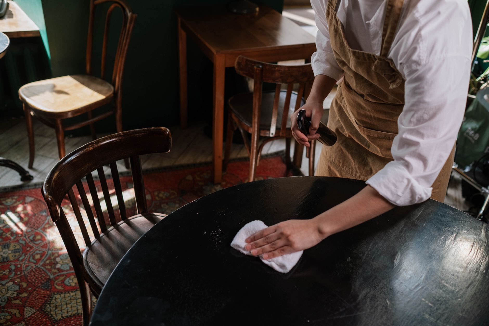 A waitress cleans a table in a cozy café with elegant wooden chairs.