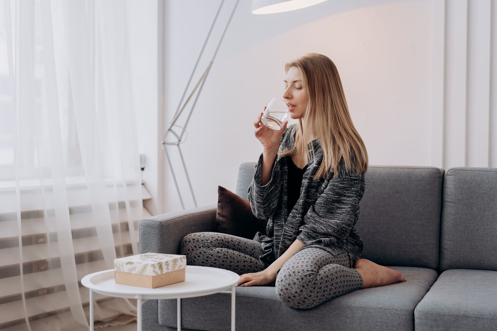 Serene woman sitting on a couch, sipping water in a cozy indoor setting.